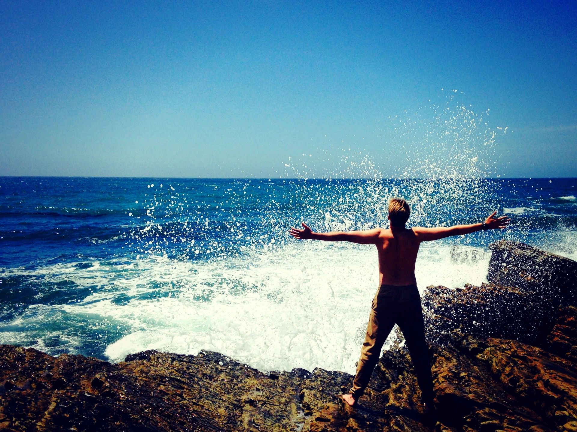 Man standing on a rocky shoreline as waves crash around him, representing resilience and growth through Crucible Therapy."